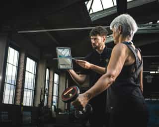 Trainer von PhysioSport erläutert Patientin am Tablet die Kraftübungen mit Kurzhanteln.