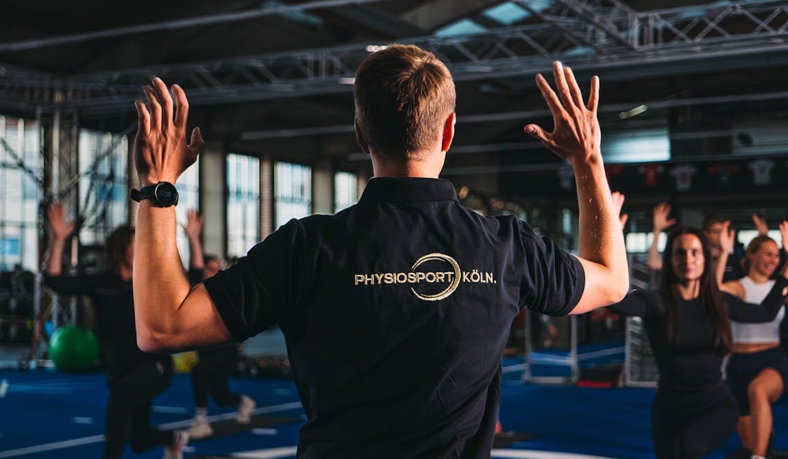 Zu sehen ist im Vordergrund ein Mitarbeiter von PhysioSport Köln von hinten, der einer Gruppe von Patienten eine Übung vormacht. Im Hintergrund eine Gruppe von Patienten.