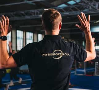 Zu sehen ist im Vordergrund ein Mitarbeiter von PhysioSport Köln von hinten, der einer Gruppe von Patienten eine Übung vormacht. Im Hintergrund eine Gruppe von Patienten.