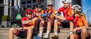 Fünf Radfahrer:innen im Sportoutfit sitzen fröhlich und entspannt auf einer Treppe und stoßen mit Mühlen Kölsch Freibier, alkoholfrei, den Partnern von Physiosport Köln, auf die beendete Radtour an.