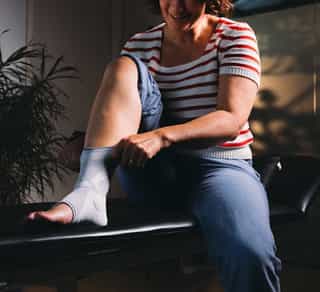 Patientin sitzt auf einer Liege bei PhysioSport Köln und begutachtet eine Sprunggelenksbandage an ihrem Fuß.