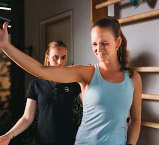Sportlerin steht mit Physiotherapeutin von PhysioSport Köln in einem Trainingsraum und hebt den rechten Arm zur Begutachtung.