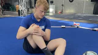 Man sieht einen Sportler bei PhysioSport Köln, schräg von vorne gesehen und sitzend am Boden, der sich vor Schmerzen an sein Knie fasst.